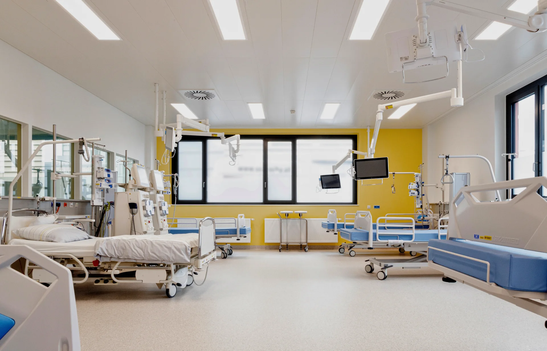 Heller, moderner Krankenhausraum mit mehreren leeren Pflegebetten, medizinischen Geräten an den Betten, Deckenlampen und Monitorarmen. Große Fenster lassen Tageslicht herein; die Wände sind gelb gestrichen, der Boden hell. Keine Personen im Raum.