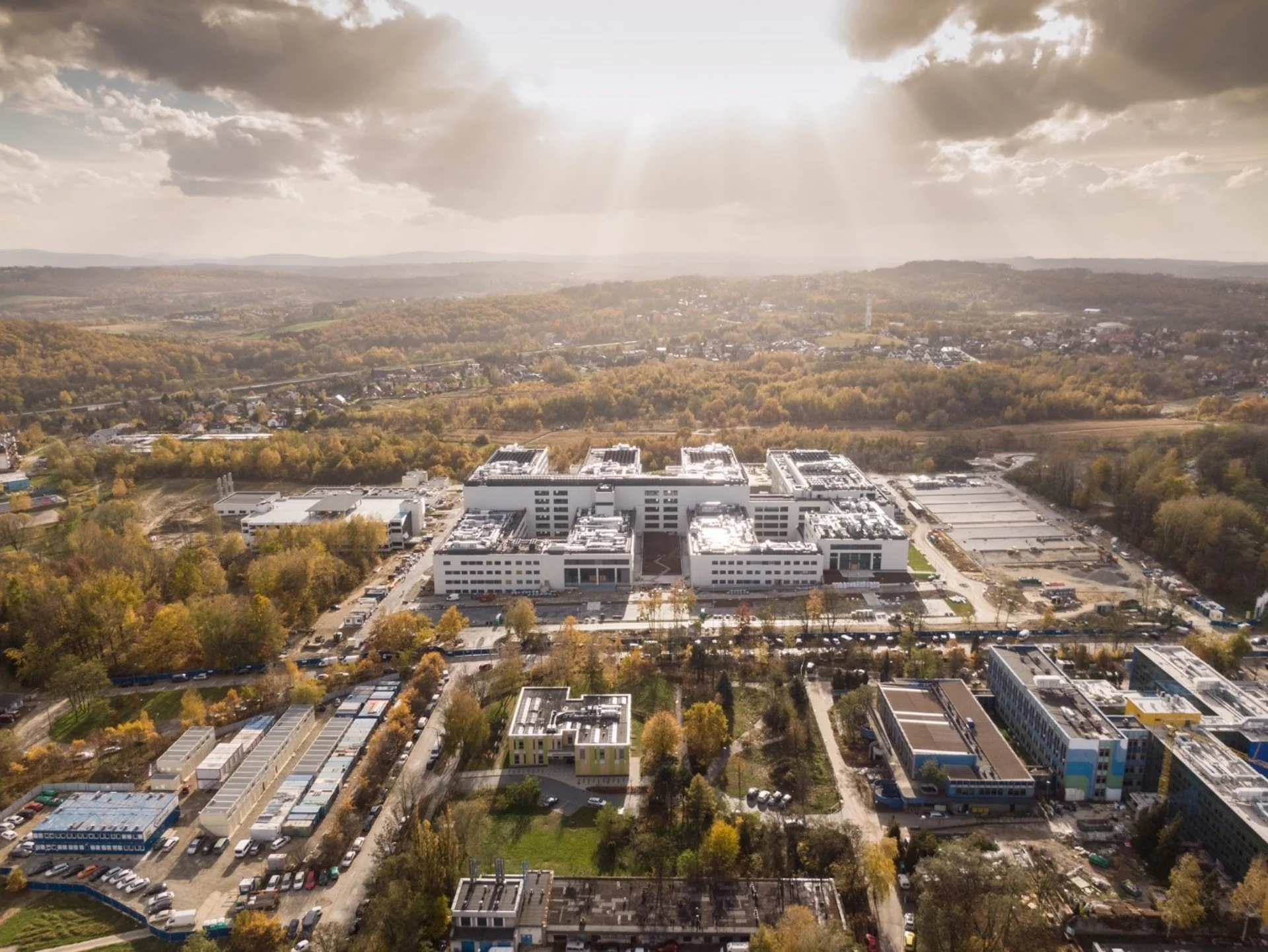 Luftaufnahme eines großen, modernen Gebäudekomplexes mit mehreren weißen, rechteckigen Gebäudeteilen und technischen Anlagen auf den Dächern. Das Gelände, auf dem sich auch noch in Bau befindliche Parkplätze befinden, ist von herbstlich gefärbten Bäumen umgeben, im Hintergrund sind Waldflächen und eine Wohnsiedlung zu sehen.